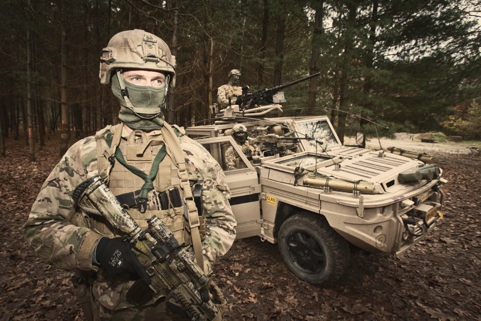 Soldiers in tactical gear beside a military vehicle in woodland